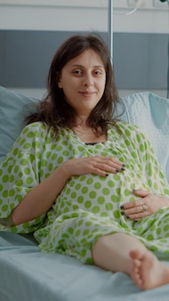 Portrait of pregnant caucasian woman sitting in hospital ward bed at healthcare clinic. impatient person with pregnancy baby bump preparing for child delivery and medical assistance