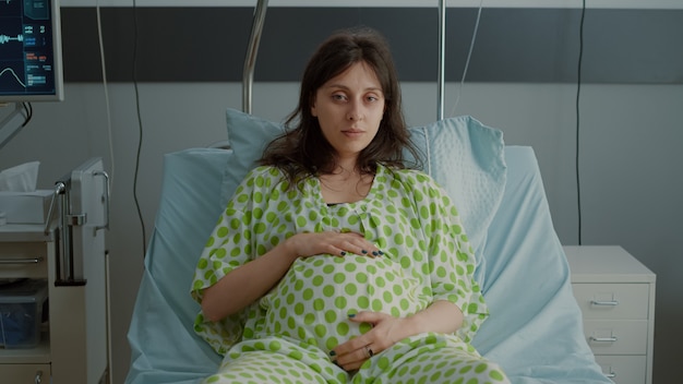 産科での出産の準備をしている医療機器を備えた病棟のベッドに横たわっている妊婦の肖像画。赤ちゃんを期待して座っている間腹に手を保持している白人患者