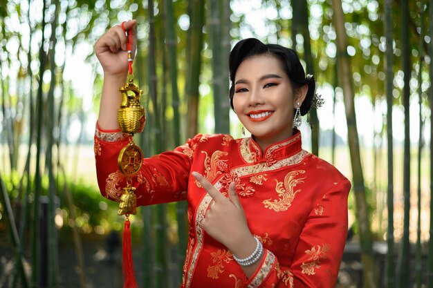中国のチャイナドレスの笑顔と中国の旧正月の幸運のシンボルの装飾のための黄金のお守りを保持している肖像画のかわいいアジアの女性