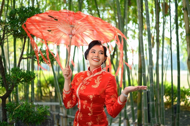 Бесплатное фото Портрет красивой азиатской женщины в китайском чонсаме, позирующей с красивым красным бумажным зонтиком в бамбуковом лесу