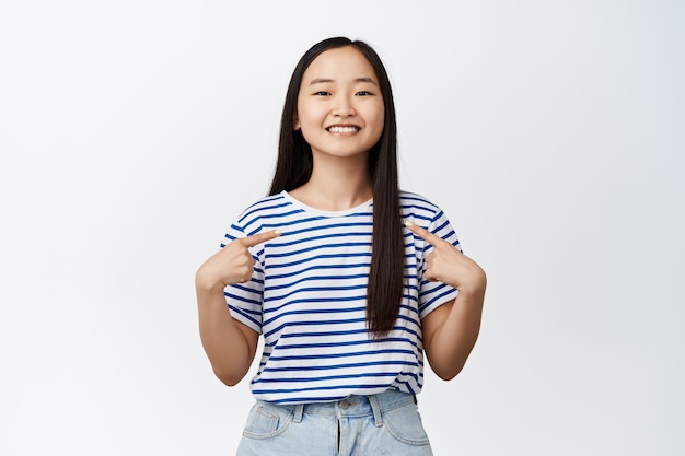 Portrait of pretty asian woman pointing fingers at herself and smiling pleased on white