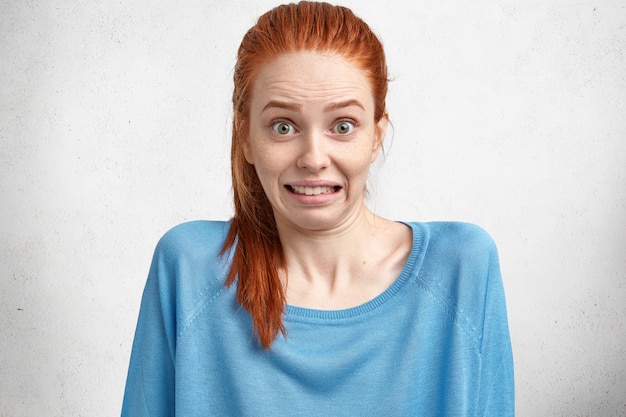 Free photo portrait of pretty beautiful redhead woman stares in bewilderment and clenches teeth, has puzzled and surprised expression