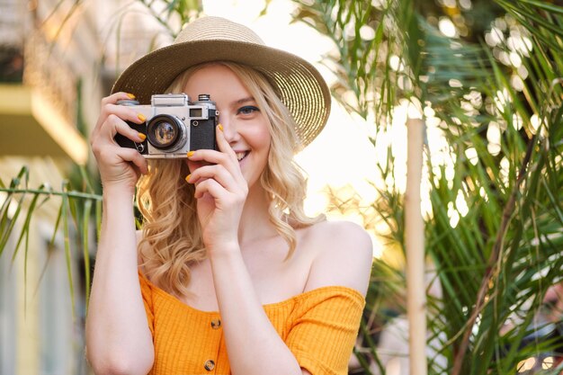Портрет симпатичной блондинки в шляпе, счастливо фотографирующей на ретро-камеру на городской улице