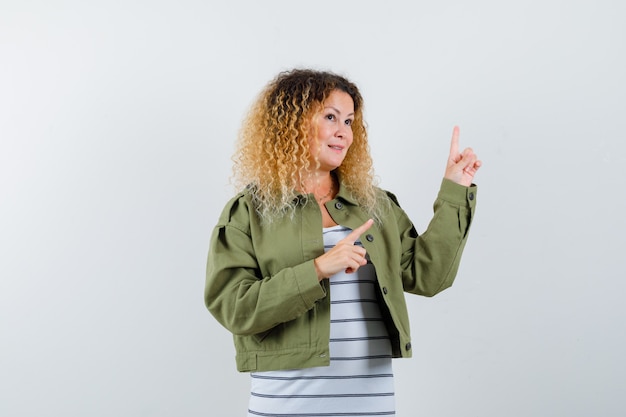 Free photo portrait of pretty blond woman pointing at upper right corner in green jacket and looking thoughtful front view