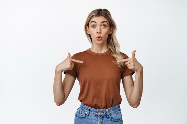 Portrait of pretty blond woman points at herself with surprised face and raised eyebrows being chosen or named standing over white background