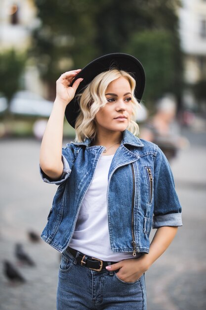 Портрет красивой блондинки в центре города, одетой в джинсовый люкс, белую футболку и черную шляпу