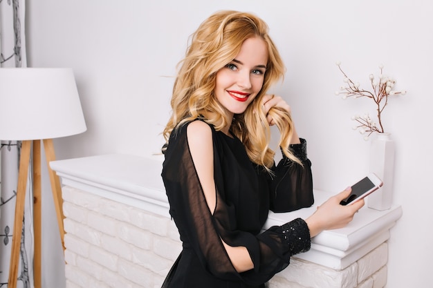 Portrait of pretty blonde girl with smartphone in hands in bright room with nice, modern white interior, standing against fake fireplace, looking. Wearing glamurus black dress.