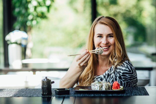 Portrait of pretty blonde lady sits in the cafe on summer terrace with sushi rolls set, after her job.