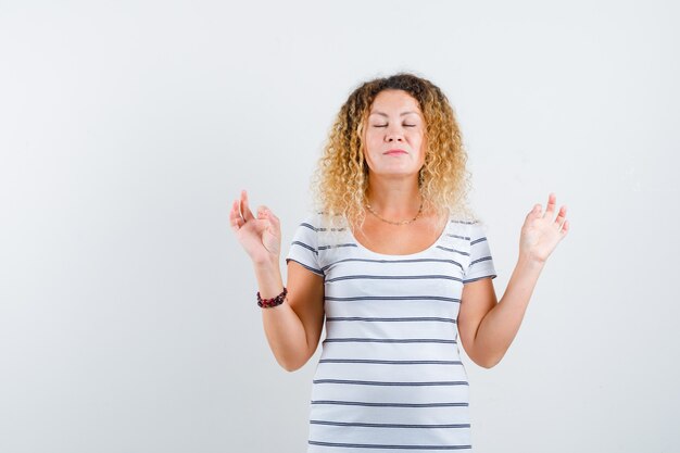 Portrait of pretty blonde woman showing yoga gesture with shut eyes in striped t-shirt and looking peaceful front view