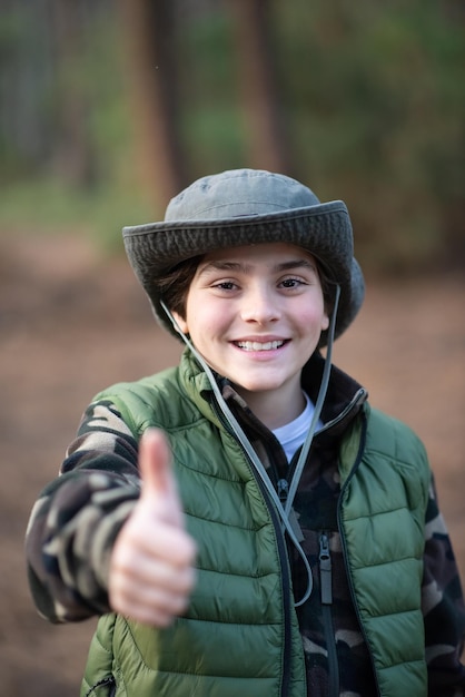 Portrait of pretty boy in panama in forest. Dark-haired schoolboy in coat smiling at camera, showing thumb up. Childhood, nature, leisure concept