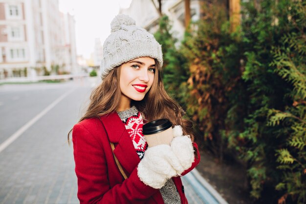 Portrait pretty brunette girl with long hair in red coat walking on street.  She holds coffee to go in white gloves, smiling .