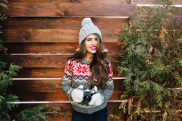 Free photo portrait pretty brunette girl with red lips in knitted hat and gloves holding camera on wooden  surround green branches.