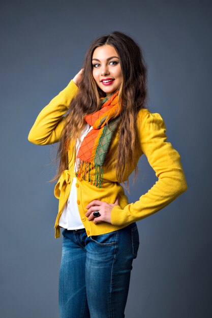 portrait of pretty brunette woman, with a long hair, bright make up, stylish cozy sweater and scarf, playful mood, gray wall, toned colors.