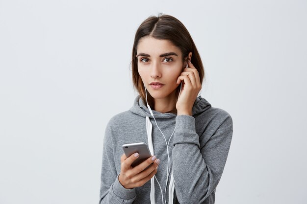 Portrait of pretty charming caucasian girl with dark long hair in stylish gray hoodie wearing earphones, searching for music to listen on smartphone,  with calm face expression.