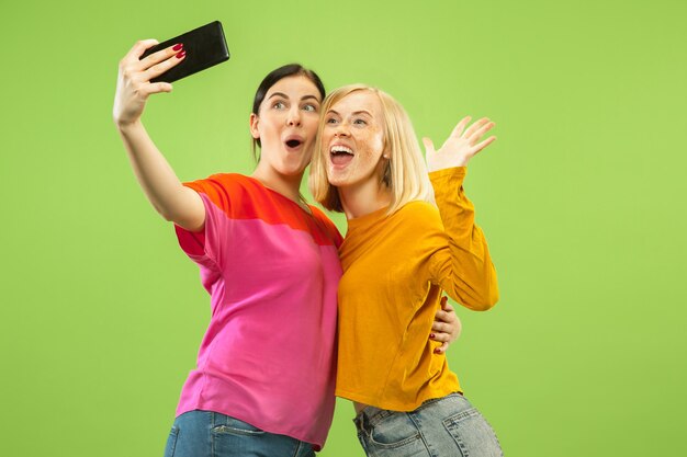 Portrait of pretty charming girls in casual outfits isolated on green studio background. Girlfriends or lesbians making selfie. Concept of LGBT, equality, human emotions, love, relation.