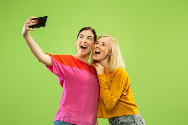 Portrait of pretty charming girls in casual outfits isolated on green studio background. Girlfriends or lesbians making selfie. Concept of LGBT, equality, human emotions, love, relation.