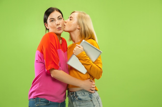 Portrait of pretty charming girls in casual outfits isolated on green studio background. Girlfriends or lesbians using a tablet for fun or payments. Concept of LGBT, human emotions, love, relation.