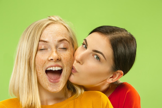 Free photo portrait of pretty charming girls in casual outfits isolated on green studio background. two female models as a girlfriends or lesbians. concept of lgbt, equality, human emotions, love, relation.