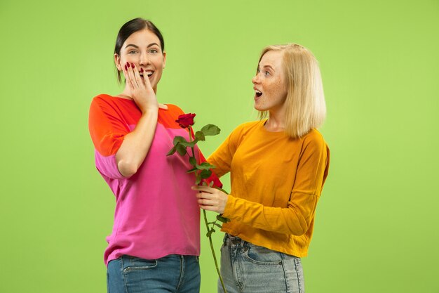 Portrait of pretty charming girls in casual outfits isolated on green studio background. Two female models as a girlfriends or lesbians. Concept of LGBT, equality, human emotions, love, relation.