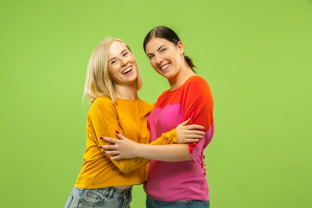 Portrait of pretty charming girls in casual outfits isolated on green studio background. Two female models as a girlfriends or lesbians. Concept of LGBT, equality, human emotions, love, relation.