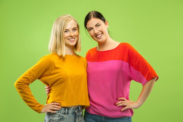 Portrait of pretty charming girls in casual outfits isolated on green studio background. Two female models as a girlfriends or lesbians. Concept of LGBT, equality, human emotions, love, relation.