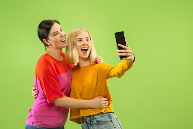 Portrait of pretty charming girls in casual outfits isolated on green wall. Girlfriends or lesbians making selfie. Concept of LGBT, equality, human emotions, love, relation.