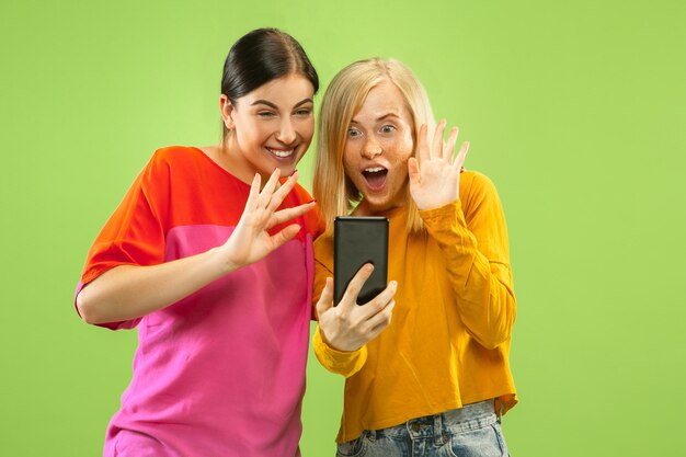 Portrait of pretty charming girls in casual outfits isolated on green wall. Girlfriends or lesbians making selfie. Concept of LGBT, equality, human emotions, love, relation.