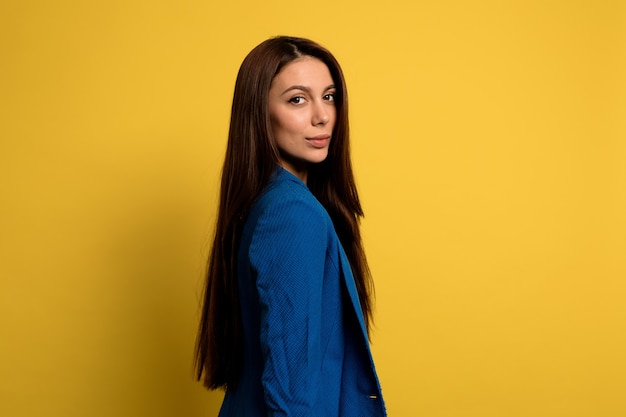 portrait of pretty charming lady with long dark hair wearing blue jacket over yellow wall