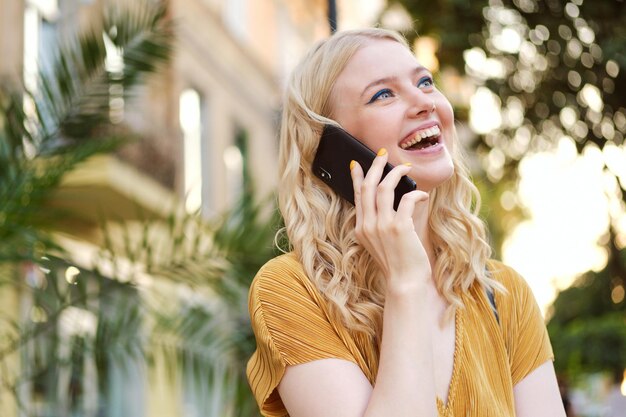 Portrait of pretty cheerful blond girl joyfully looking away while talking on cellphone on city street