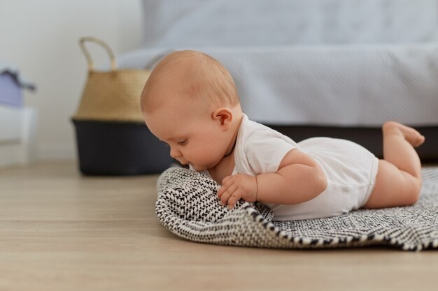 Portrait of pretty crawling baby girl posing indoor of floor, studying world around, small kid wearing white bodysuit, lying on grey carpet near sofa.