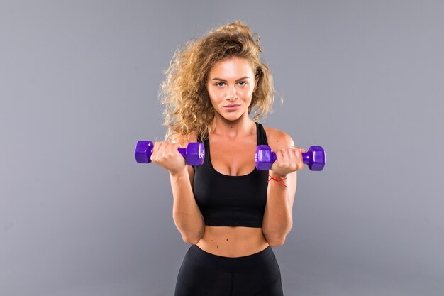 Portrait of pretty curly sporty girl holding weights dumbbels isolated on gray wall