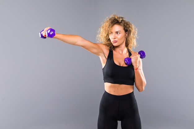 Portrait of pretty curly sporty girl holding weights dumbbels isolated on gray wall