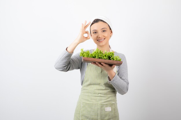 Portrait of a pretty cute woman with a wooden board of fresh lettuce showing ok gesture