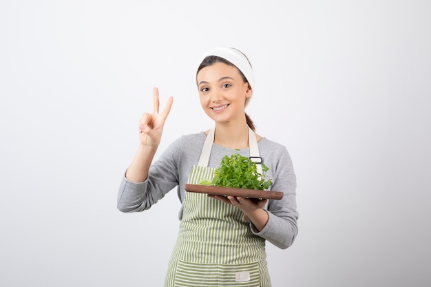 Portrait of a pretty cute woman with a wooden board of fresh parsley showing victory sign