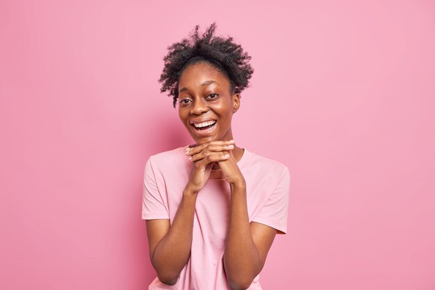 Portrait of pretty dark skinned Afro American woman keeps hands under chin smiles broadly 