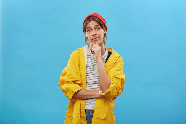 Portrait of pretty female dressed in yellow raincoat and red hat keeping her hand on chin looking directly going to have walk in forest or to river isolated