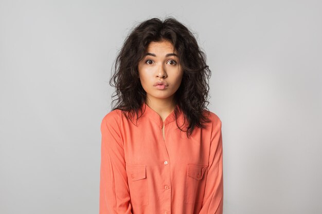 Portrait of pretty funny woman with shocked expression of face, curly hair, natural make-up, young hipster, surprised emotion, isolated, orange blouse