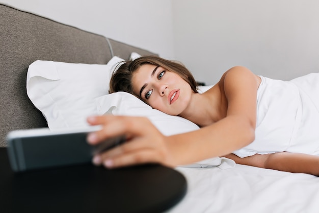 Portrait pretty girl on bed in modern apartment. She looks at phone on table.