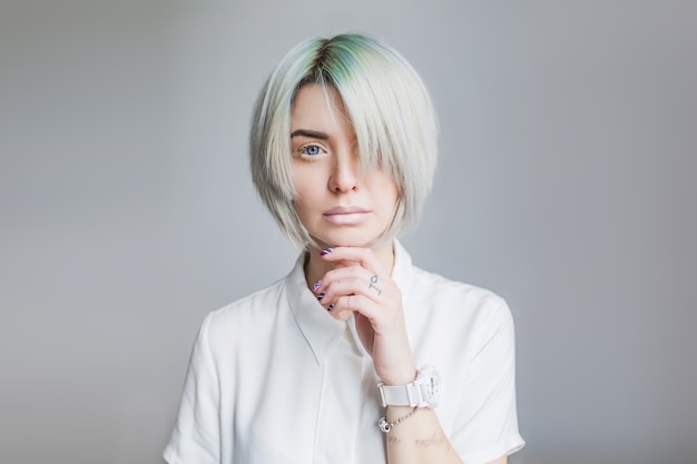 Free photo portrait of pretty girl with gray short hairstyle on gray background. she wears white dress and light makeup. bangs covers half the face. her hand touches face.