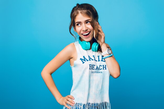 Portrait of pretty girl with long curly hair in tail talking on phone on blue background in studio. She wears white T-shirt, blue headphones on shoulders.