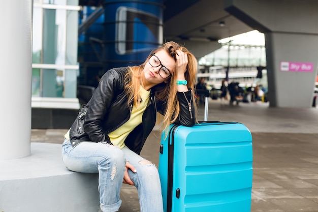 Портрет красивой девушки с длинными волосами в очках, сидящих снаружи в аэропорту. Она носит желтый свитер с черной курткой и джинсами. Она прислонилась к чемодану, и ей скучно ждать.