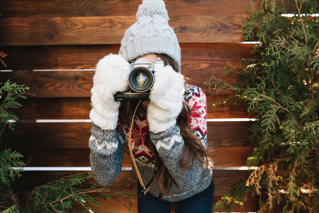 Portrait pretty girl with long hair in knitted hat and gloves making a photo on camera on wooden .