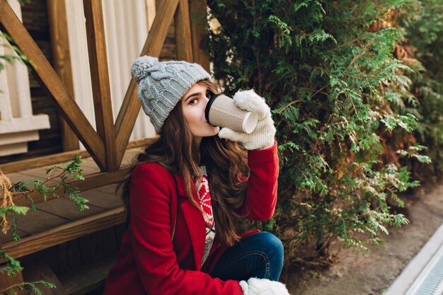 Portrait pretty girl with long hair in red coat, knitted hat and white gloves sitting on wooden stairs outdoor. She drinks coffee and looking .