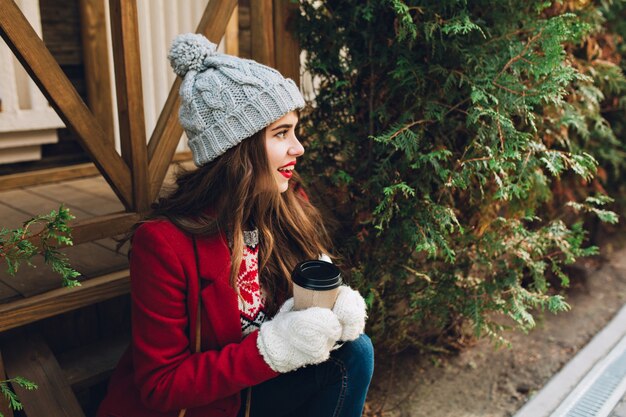 Портрет красивой девушки с длинными волосами в красном пальто, сидя на деревянной лестнице на открытом воздухе. На ней серая вязаная шапка, белые перчатки, она держит кофе и улыбается в сторону.