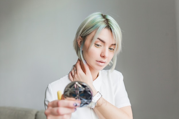 Portrait of pretty girl with short gray  hairstyle on gray background. She wears white dress and looks to the mirror in hand.