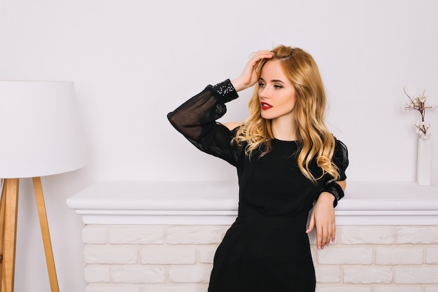 Portrait of pretty girl, young woman with blonde wavy hair sensually looking to side touching her hair. Wearing stylish black dress. White wall, fireplace, lamp.