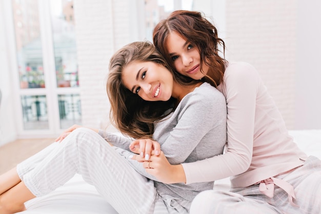 Free photo portrait of pretty girls in pajamas on bed. girl with curly hair is hugging from back other girl, they smiling to camera