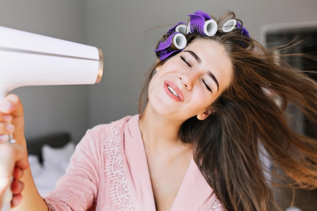 Free photo portrait pretty housewife in pink bathrobe with curler at home. she looks enjoyed at drying hair