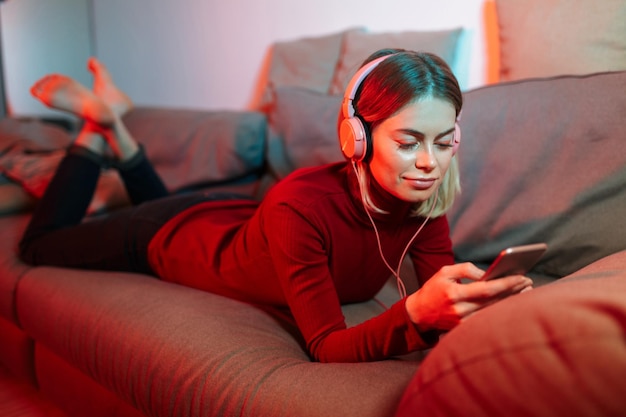 Portrait of pretty lady lying on sofa in headphones and cellphone in hands Young cool woman listening music while spending time at home