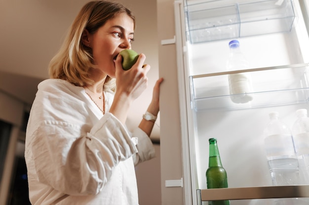 夜にキッチンに立って、家で青リンゴを食べながらオープン冷蔵庫で見ているきれいな女性の肖像画